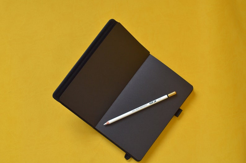 Pu&ograve; includere: Un taccuino nero con una matita bianca aperta su una superficie gialla.