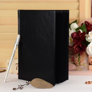 May include: A black leather-bound guest book with a white pen lying on a white surface. The book is closed and has a smooth, textured cover. There are flowers and a wooden sign with the word "August" in the background.