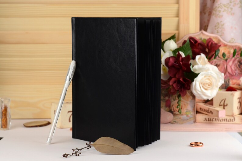 May include: A black leather-bound guest book with a white pen lying on a white surface. The book is closed and has a smooth, textured cover. There are flowers and a wooden sign with the word "August" in the background.
