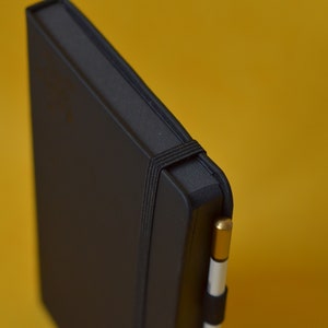 Pu&ograve; includere: Un taccuino nero rilegato in pelle con chiusura a elastico nero e una penna oro e bianca infilata nella fascia.