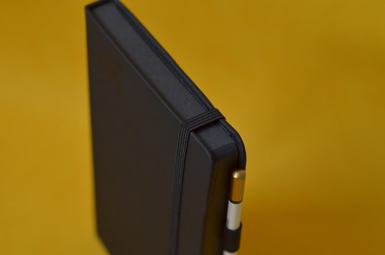 Pu&ograve; includere: Un taccuino nero rilegato in pelle con chiusura a elastico nero e una penna oro e bianca infilata nella fascia.