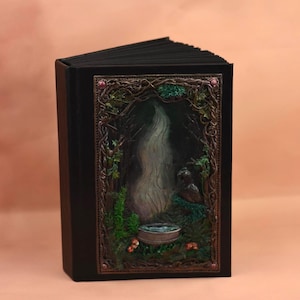 May include: A black journal with a detailed forest scene on the cover. The cover art features a waterfall, a small pool, and a detailed frame of vines and leaves. The journal is open, revealing the black pages inside.