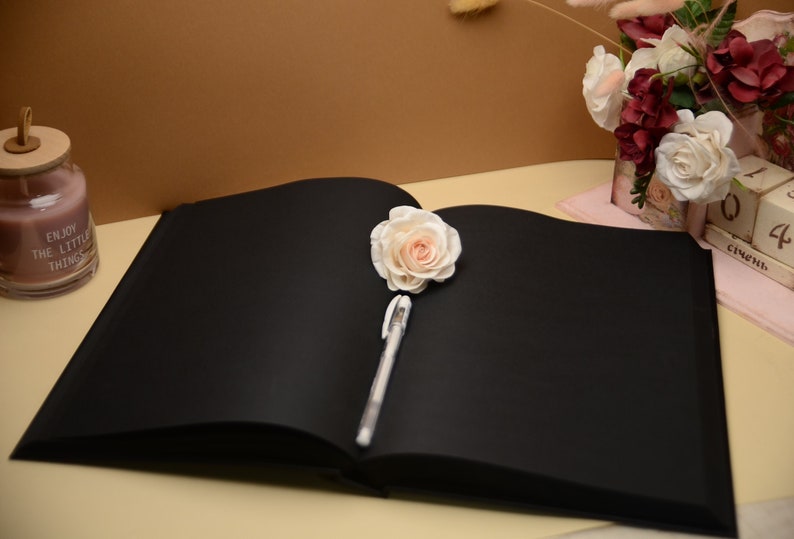 Puede incluir: Un diario de tapa dura negro con una pluma blanca y una rosa blanca encima de las p&aacute;ginas. El diario est&aacute; abierto y las p&aacute;ginas est&aacute;n en blanco. El diario est&aacute; sobre una superficie marr&oacute;n claro.