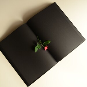 Puede incluir: Un libro negro con una sola rosa roja y hojas verdes en las p&aacute;ginas abiertas.
