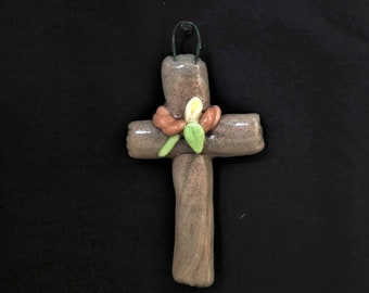 Op de afbeelding: Een bruin klei kruis met een groene blad- en een oranje bloemenversiering. Het kruis hangt aan een draadlus.