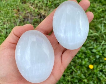 May include: Two white selenite palm stones, also known as satin spar, with a smooth, polished surface and a striated pattern. The stones are oval-shaped and have a translucent appearance.