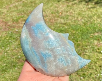 May include: A crescent moon shaped carving made of blue and gray agate. The stone has a swirling pattern of blue and gray.