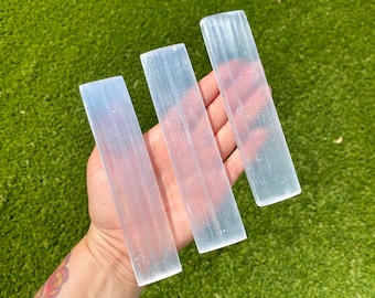 May include: Three selenite wands, also known as satin spar, are shown in a hand. The wands are clear and translucent with striations. The wands are approximately 4 inches long.