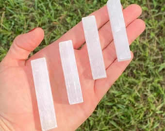 May include: Four white selenite sticks of varying sizes, arranged in a row on a hand. The sticks are smooth and have a natural, striated texture.