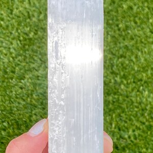 May include: A white selenite crystal wand with a smooth, polished surface. The wand is held in a hand against a green background.