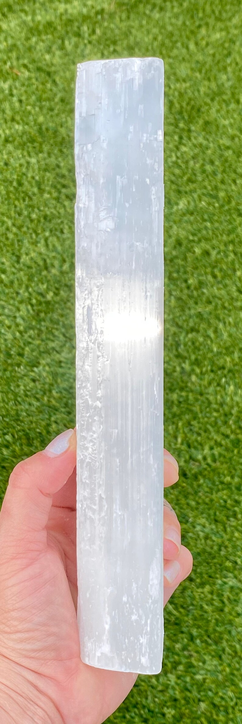 May include: A white selenite crystal wand with a smooth, polished surface. The wand is held in a hand against a green background.