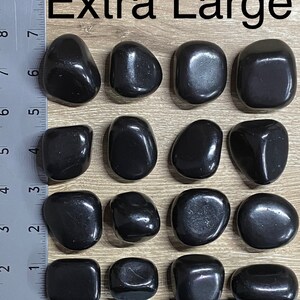 May include: Extra Large black polished stones. The stones are smooth and vary in shape, with rounded and rectangular forms. The image includes a ruler for size reference. The text "Extra Large" is at the top.