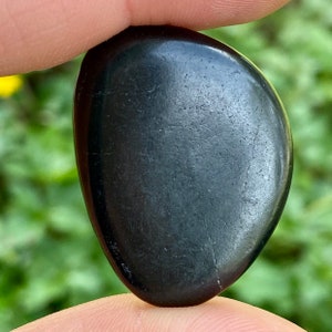 May include: A smooth, black, polished stone with a rounded shape. The stone is held between two fingers, with a blurred green background. The stone has a glossy finish and appears to be a natural material.