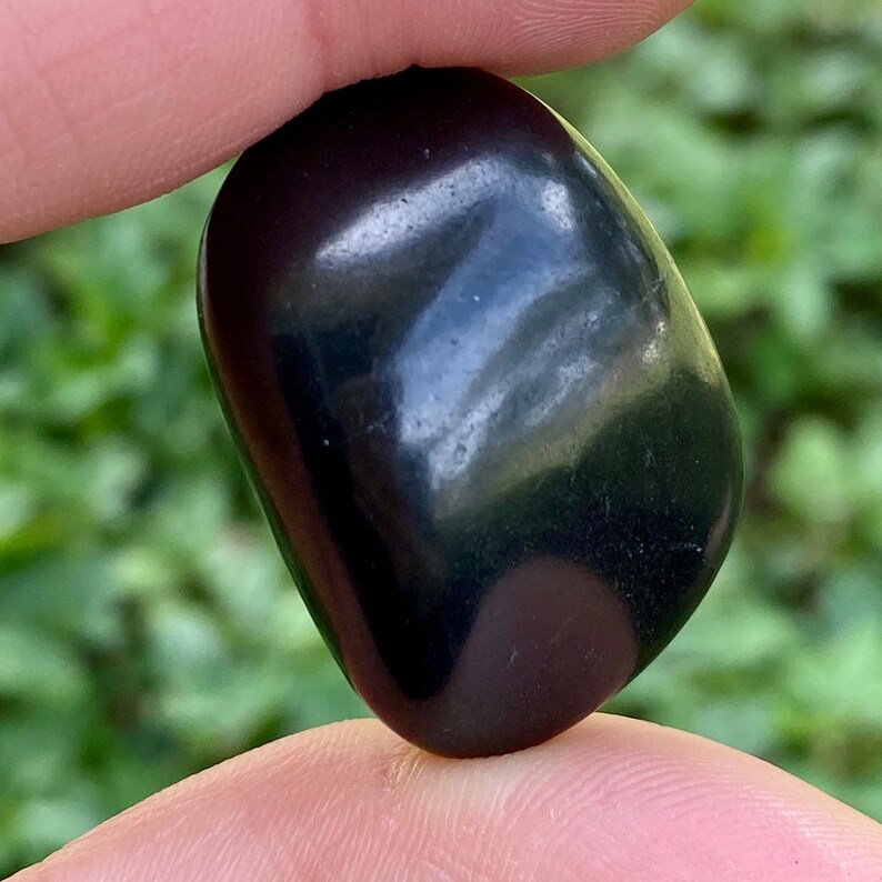 May include: A polished, dark black stone with a smooth, rounded shape. The stone is held between two fingers, with a blurred green background of foliage. The surface of the stone reflects light, creating highlights.