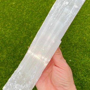 May include: A large, white selenite wand with a smooth, polished surface and visible striations. The wand is held in a hand, showcasing its size and shape.