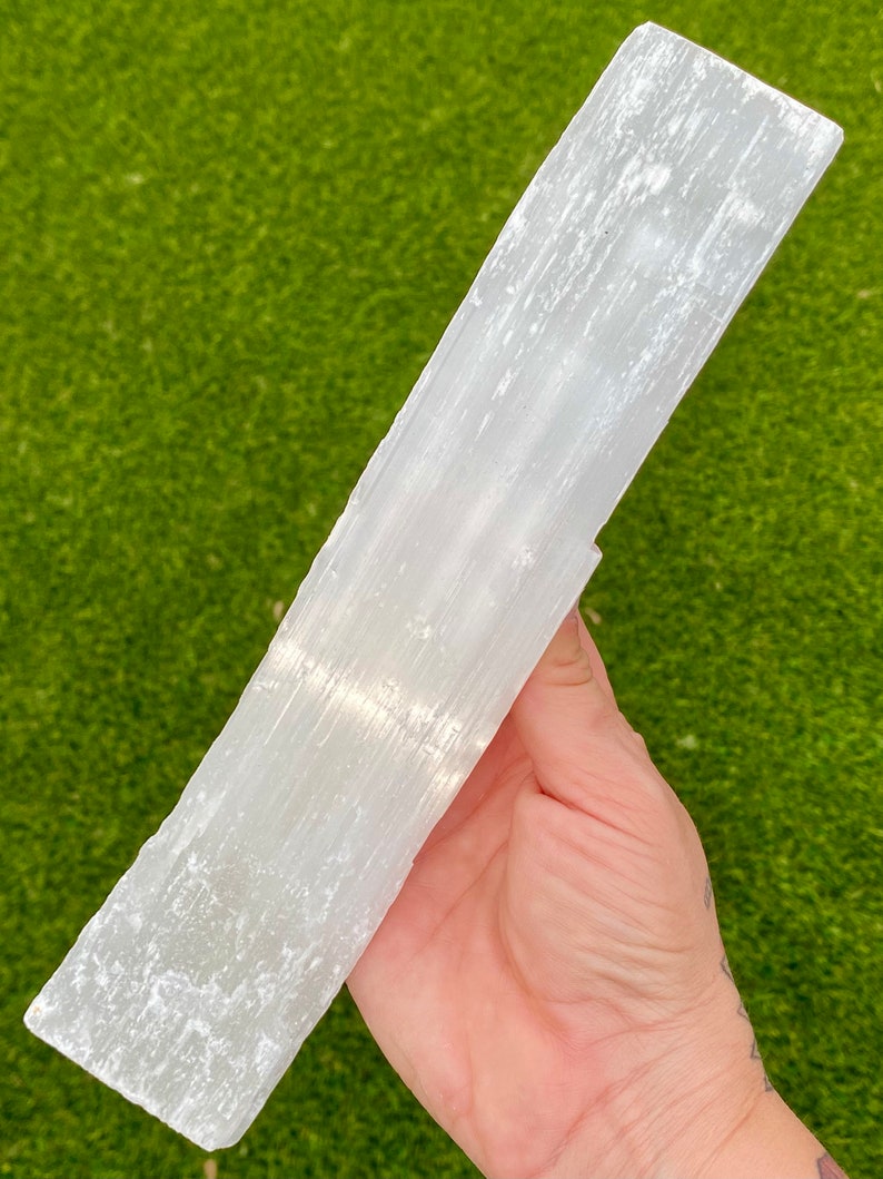 May include: A large, white selenite wand with a smooth, polished surface and visible striations. The wand is held in a hand, showcasing its size and shape.