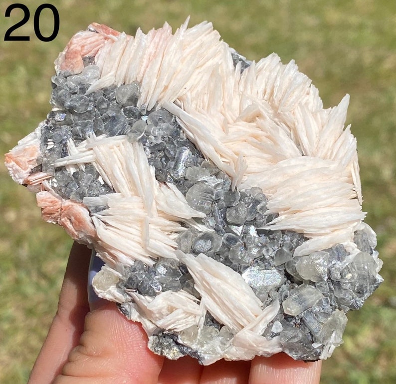May include: A cluster of white barite crystals with a pink hue, intergrown with gray and clear quartz crystals. The crystals have a layered, blade-like structure.