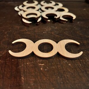 May include: A collection of wooden triple moon symbols, a Wiccan and Pagan symbol, are shown on a dark wooden surface. The light brown symbols are crescent-shaped, with a central circle. More symbols are scattered in the background.