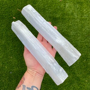 May include: Two white selenite wands, also known as selenite sticks, are held in a hand. The wands are smooth and have a striated texture.