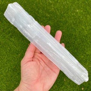 May include: A white selenite wand with a smooth, striated surface. The wand is approximately 8 inches long and is held in a hand.