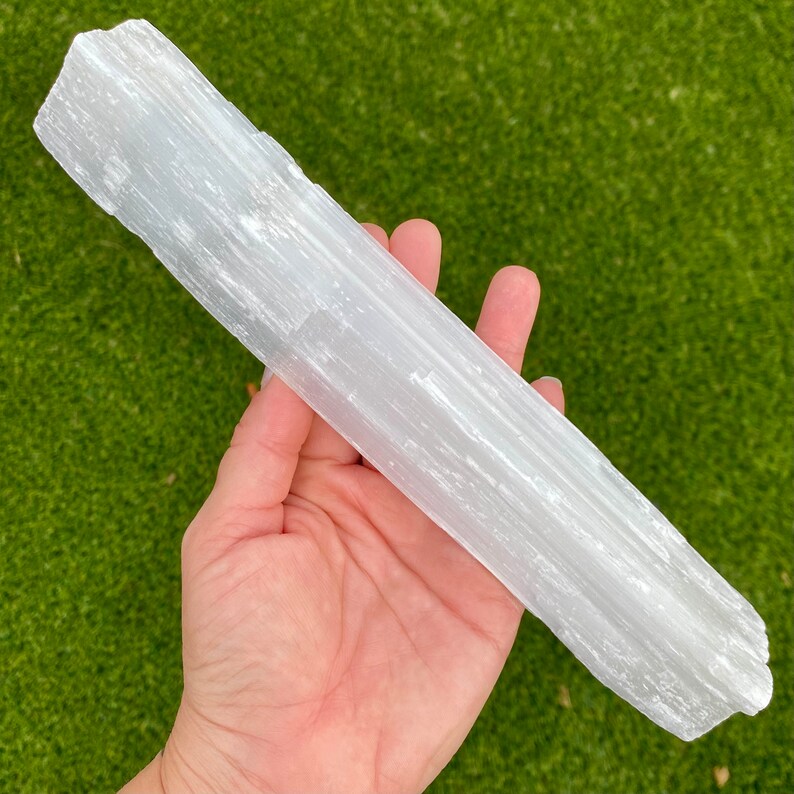May include: A white selenite wand with a smooth, striated surface. The wand is approximately 8 inches long and is held in a hand.