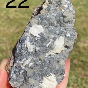 May include: A close-up of a gray and white mineral specimen with the number 22 written on the top left corner of the image. The mineral has a rough, textured surface and is covered in small, white crystals.