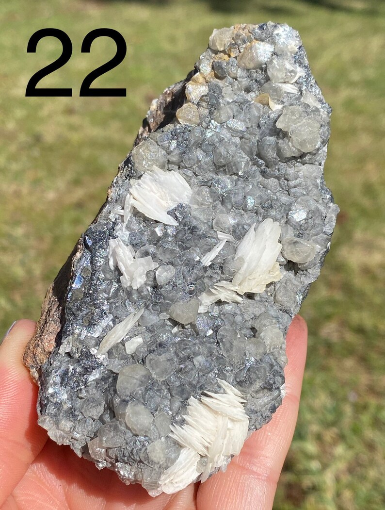 May include: A close-up of a gray and white mineral specimen with the number 22 written on the top left corner of the image. The mineral has a rough, textured surface and is covered in small, white crystals.