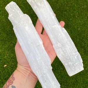 May include: Two white selenite wands, also known as selenite sticks, are held in a hand. The wands are smooth and have a striated texture.