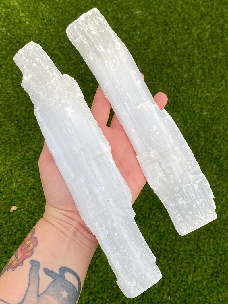 May include: Two white selenite wands, also known as selenite sticks, are held in a hand. The wands are smooth and have a striated texture.