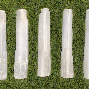 May include: Five white selenite wands, also known as selenite sticks, are arranged in a row on a green artificial grass background. The wands are smooth and have a natural, crystalline texture.