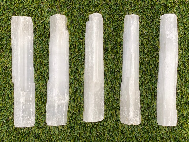 May include: Five white selenite wands, also known as selenite sticks, are arranged in a row on a green artificial grass background. The wands are smooth and have a natural, crystalline texture.