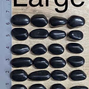 May include: Twenty-one polished black stones, labeled "Large", are arranged on a wooden surface. The stones are smooth and oval-shaped, with a glossy finish. A ruler is placed along the side for scale.
