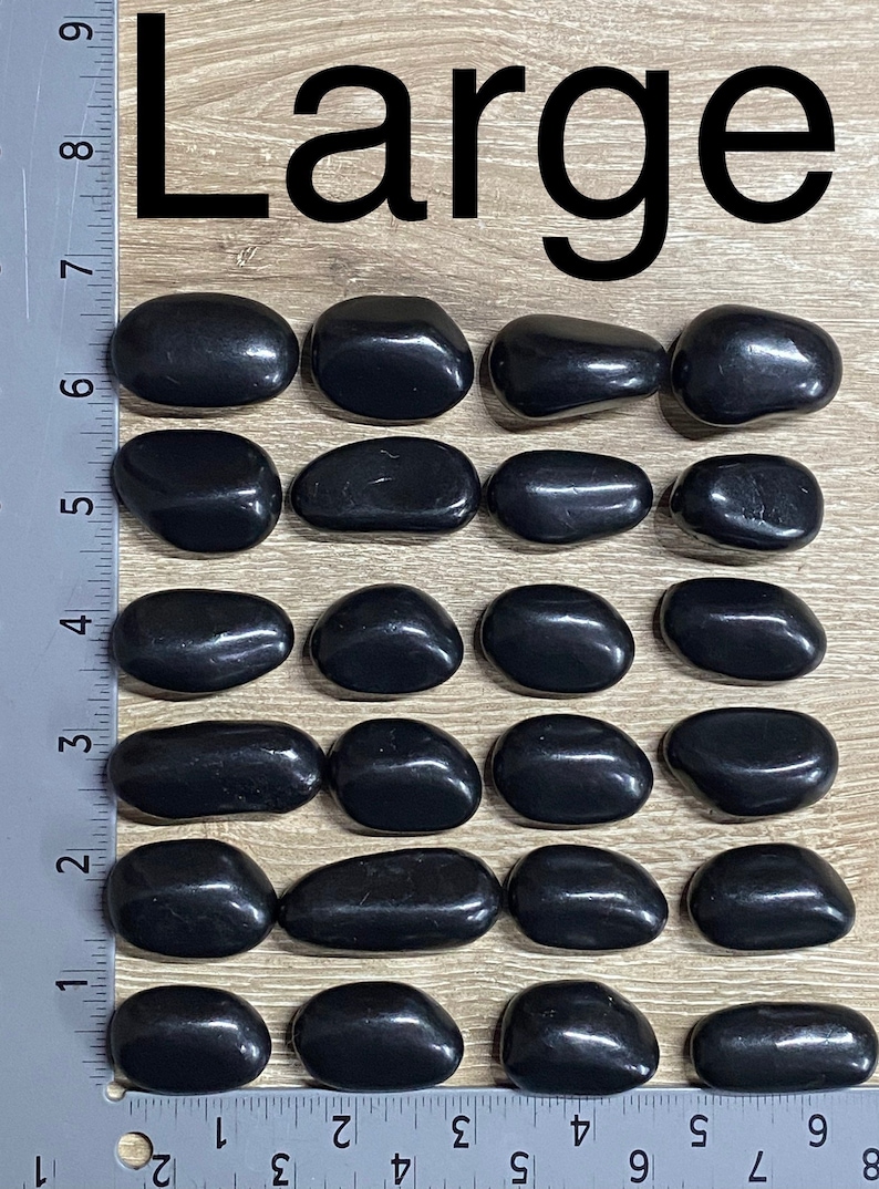 May include: Twenty-one polished black stones, labeled "Large", are arranged on a wooden surface. The stones are smooth and oval-shaped, with a glossy finish. A ruler is placed along the side for scale.