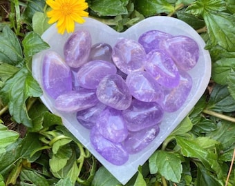 May include: A heart-shaped white dish filled with tumbled amethyst stones. The stones are a light purple color and have a smooth, polished finish.