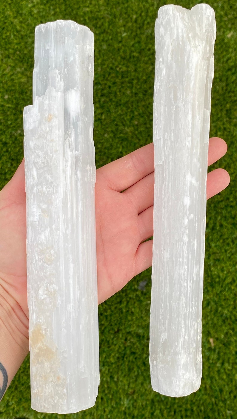 May include: Two white selenite wands, also known as selenite sticks, are held in a hand. The wands are about 6 inches long and have a smooth, polished surface.