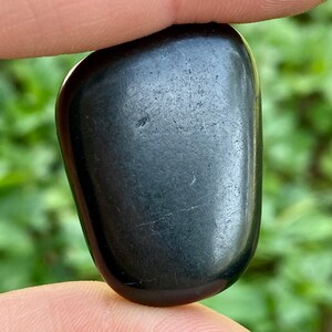 May include: A smooth, polished black stone, held between fingers, with a slightly irregular shape. The stone has a glossy surface and subtle variations in tone, suggesting natural origins. The overall appearance is dark and sleek.