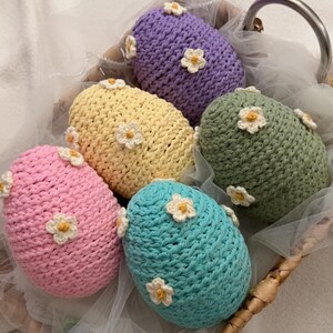 May include: A collection of six crocheted egg-shaped items in a woven basket. Each egg is a different pastel color: pink, yellow, blue, green, and purple. Small white and yellow crocheted flowers are attached to each egg.