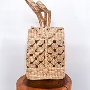 May include: A woven straw tote bag with a rectangular shape and intricate cut-out design. The bag has two long handles and a knotted closure.