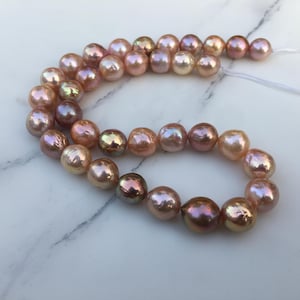 Puede incluir: Un collar de perlas de agua dulce iridiscentes en tonos rosa, melocotón y dorado. Las perlas tienen forma irregular y están ensartadas en un hilo blanco. Las perlas miden aproximadamente 0,8 cm de diámetro.