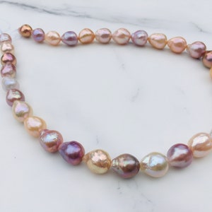 Puede incluir: Un collar de perlas de forma irregular, con colores variados, que van del rosa al lavanda y crema. Las perlas tienen superficies iridiscentes. El collar está compuesto por perlas ensartadas, creando una joya elegante.