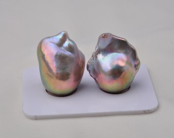 May include: Two iridescent baroque pearls with a pearlescent sheen in shades of pink, green, and blue. The pearls are displayed on a white surface.