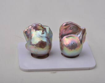 May include: Two iridescent baroque pearls with a pink and green hue, displayed on a white surface.
