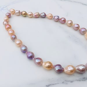 Puede incluir: Una hebra de perlas barrocas en tonos rosa, lavanda y crema. Las perlas están enhebradas en un cordón blanco y están dispuestas en un patrón graduado.