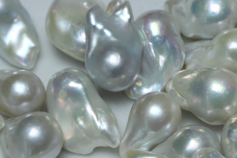 May include: A collection of white, pearlescent, baroque pearls of various shapes and sizes. The pearls have an iridescent sheen and are arranged on a white surface.