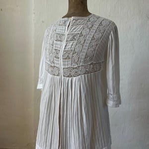 May include: A white blouse featuring vertical pleats and intricate lace panels across the chest and shoulders. The garment has three-quarter length sleeves with lace-trimmed cuffs, a round neckline, and a front button closure, displayed on a mannequin.