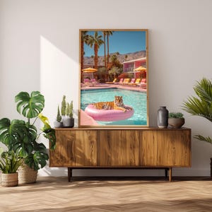 May include: A framed art print depicts a tiger lounging on a pink inflatable ring in a swimming pool. The pool scene is set against a backdrop of palm trees, a pink building, and a clear blue sky. The print is displayed on a wooden cabinet.