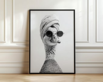 May include: Black and white framed print of a goose wearing a rolled towel on its head and round sunglasses. The goose is centered in the frame, with a neutral background. The print is displayed against a white wall and a wooden floor.