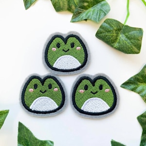 May include: Three green felt frog patches with white eyes and a white belly. The frogs have a smiling face with pink cheeks. The patches are on a white background.