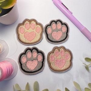 May include: Four embroidered iron-on patches in the shape of a paw print with a pink heart in the centre. Two patches are beige with a brown outline and two patches are grey with a black outline.
