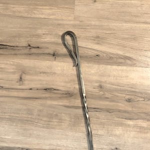May include: A hand-forged metal fire poker with a twisted shaft and a looped handle. The metal has a silver colour and a rustic appearance. The poker is designed for use in a fireplace or fire pit.